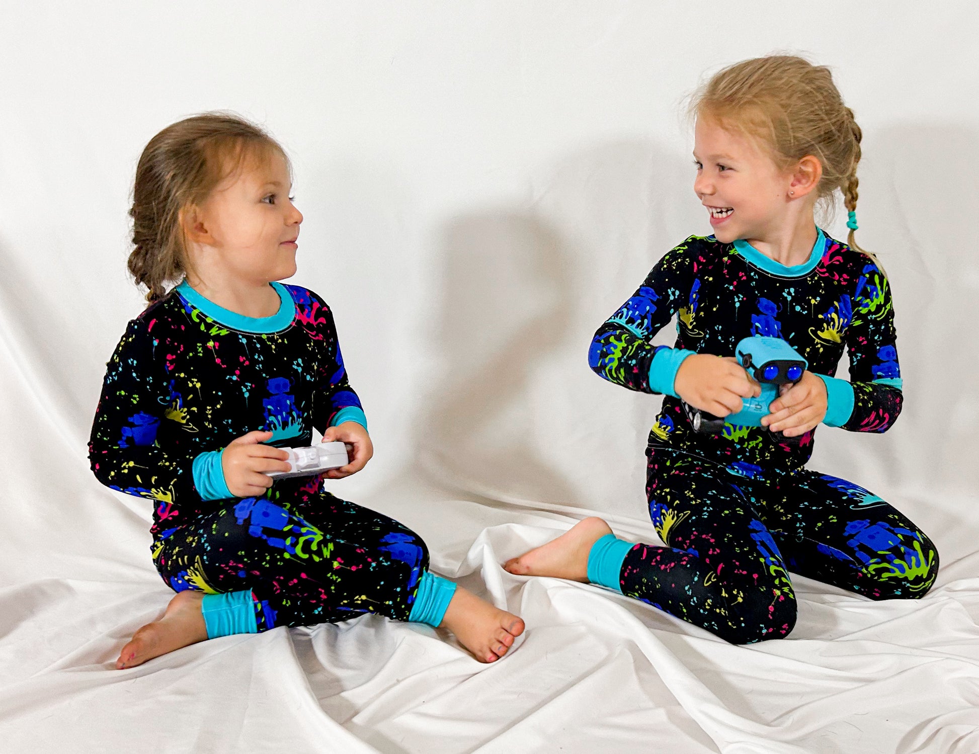 two girls playing on the floor while wearing matching pajamas dorned with neon robots and paint splashes