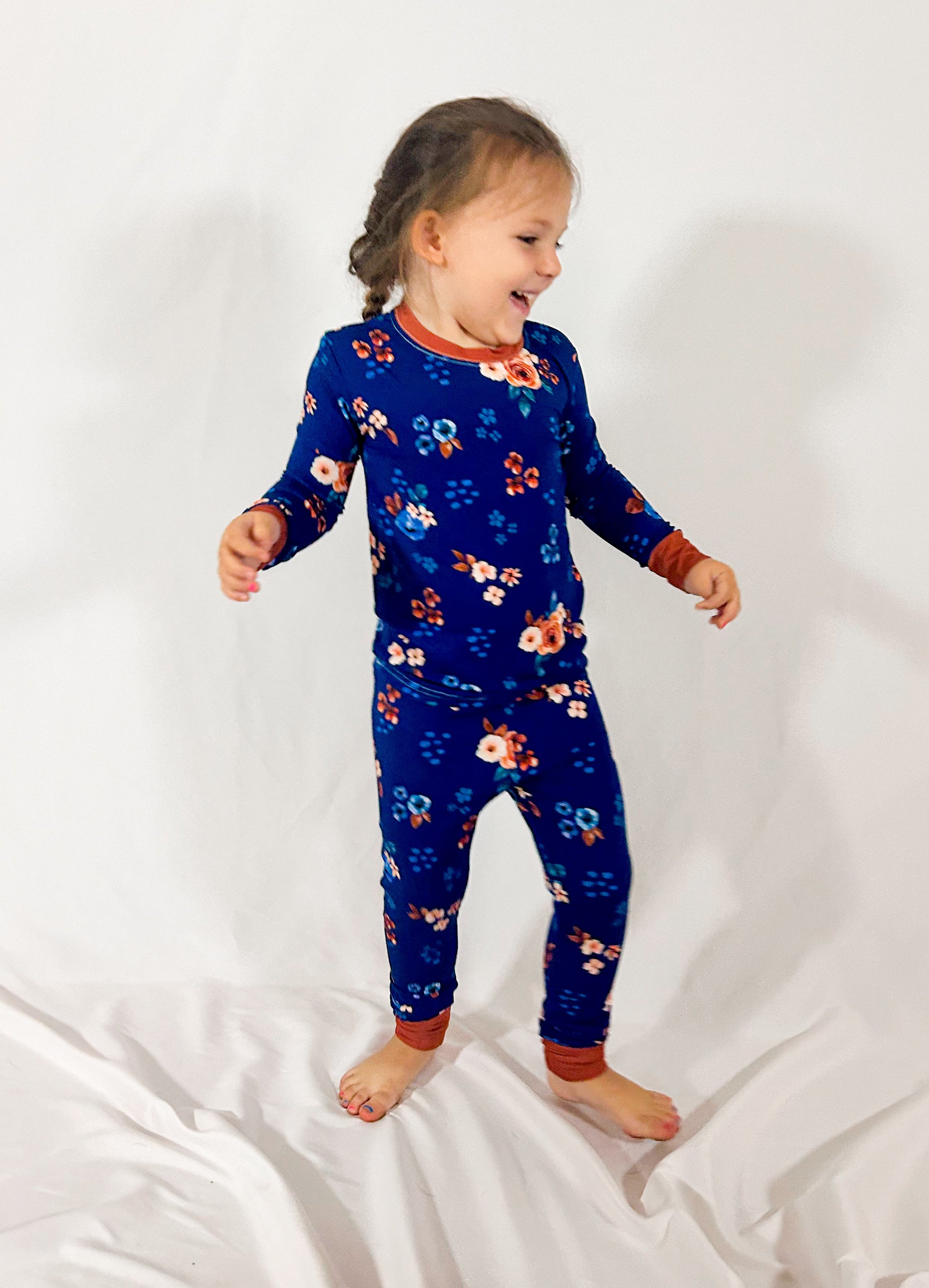 A child wearing a two-piece pajama set with a navy blue background and a floral print including roses and berries. The set has contrasting brown trim and the child is standing on a light background.