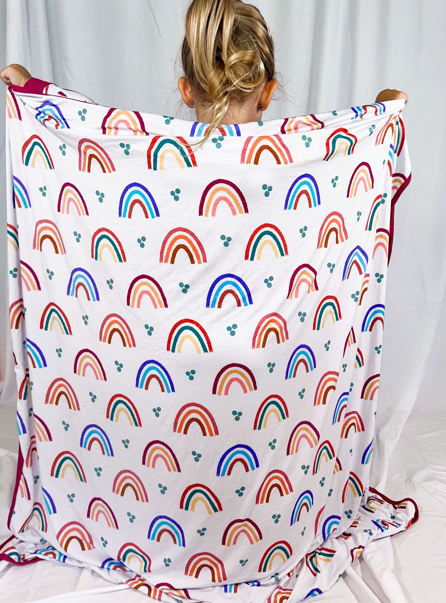 A person holding up a multicolored blanket with a geometric pattern, featuring rainbows, in front of a white background.