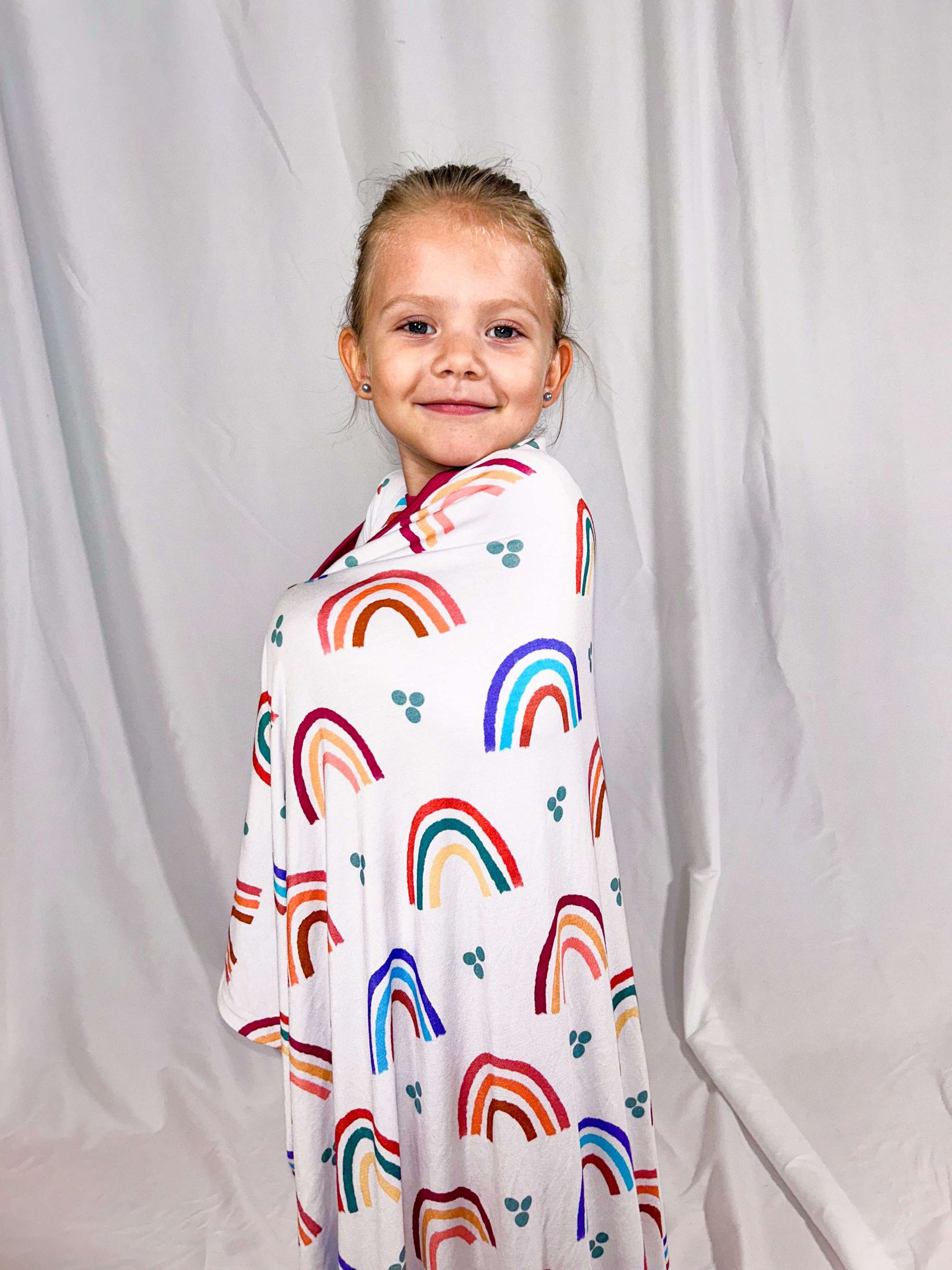A child standing and smiling, wrapped in a multicolored bamboo blanket with a rainbow pattern.