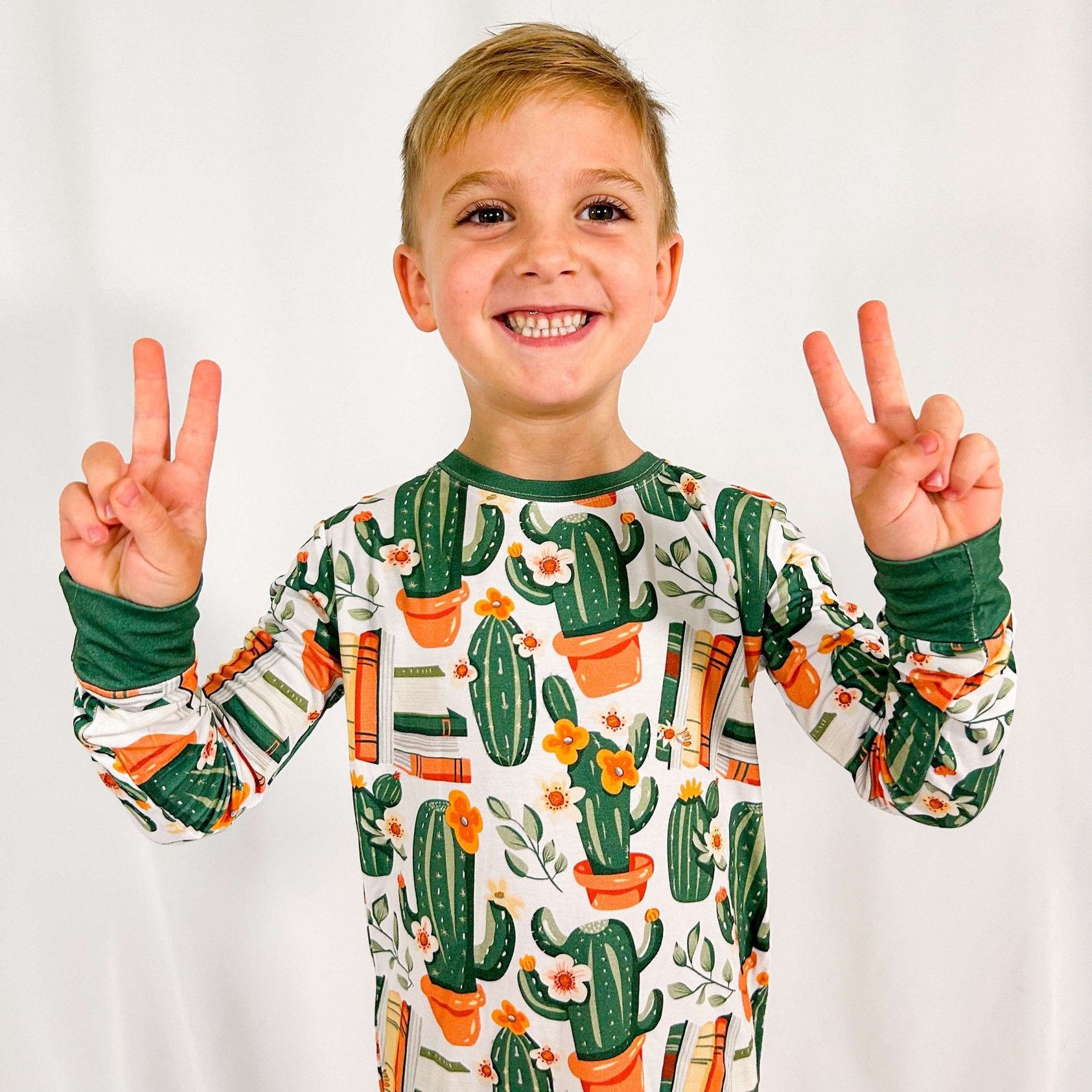 A child wearing a two-piece pajama set with a cacti and books print, smiling and making a peace sign.