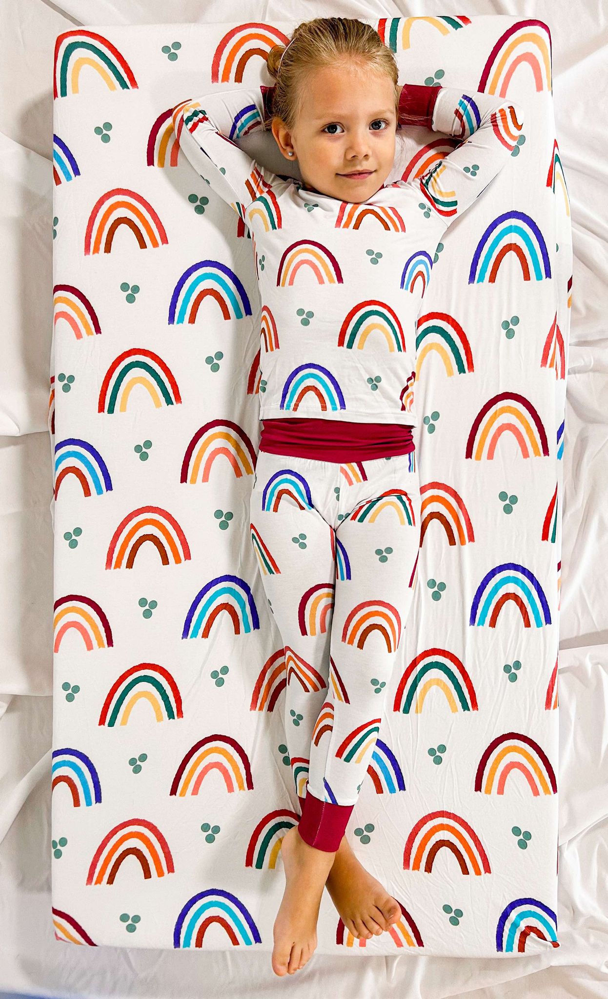 A child lying on a crib sheet with a colorful rainbow pattern.