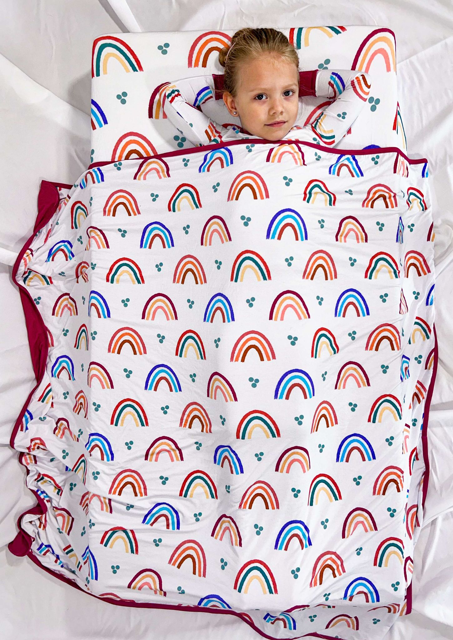 A baby crib sheet with a colorful rainbow pattern and a solid red border, the sheet is spread over a crib with a baby lying on top.