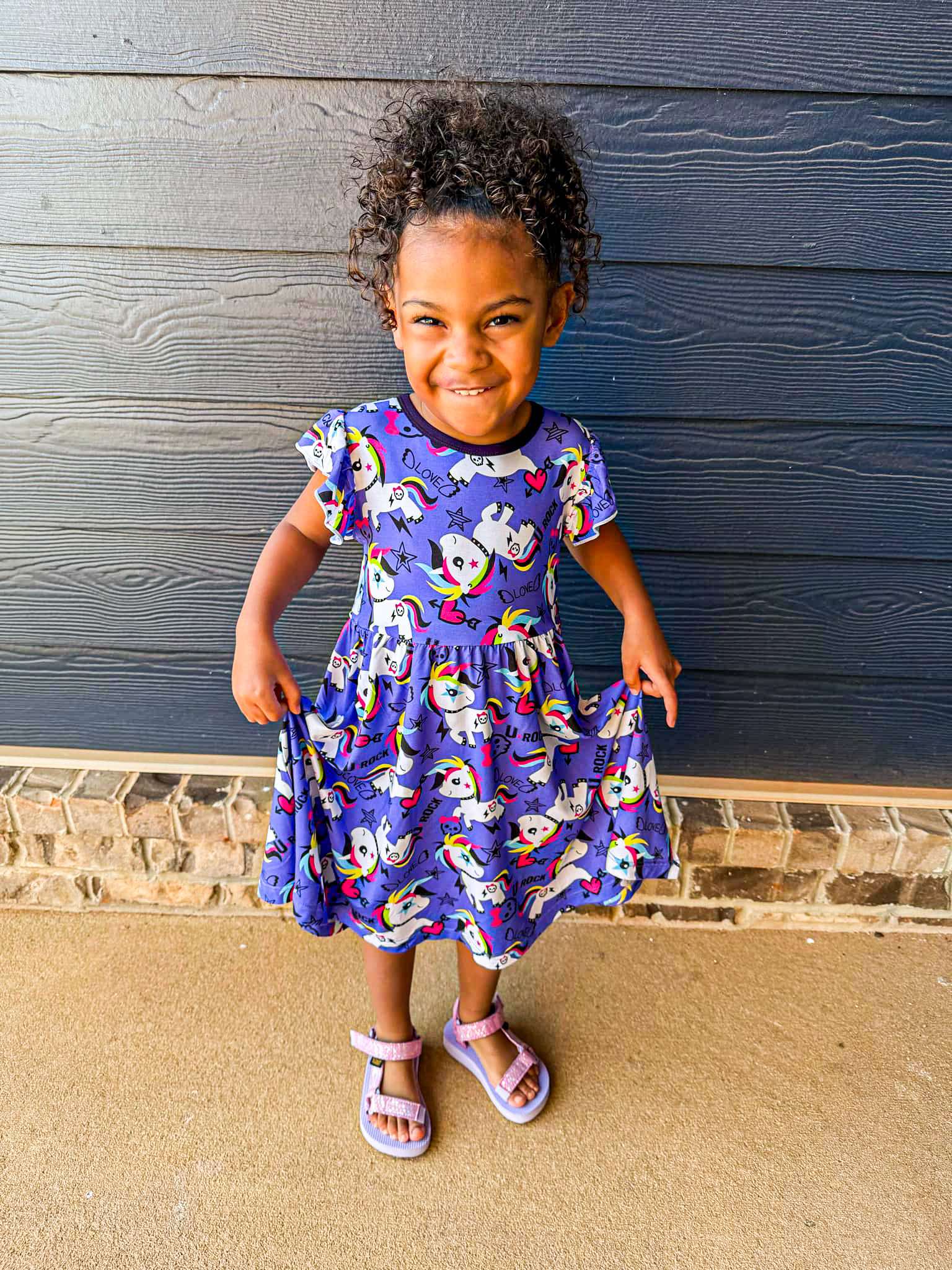 A young child standing in front of a dark wall, wearing a short-sleeved dress with a pattern of punk-inspired unicorns.