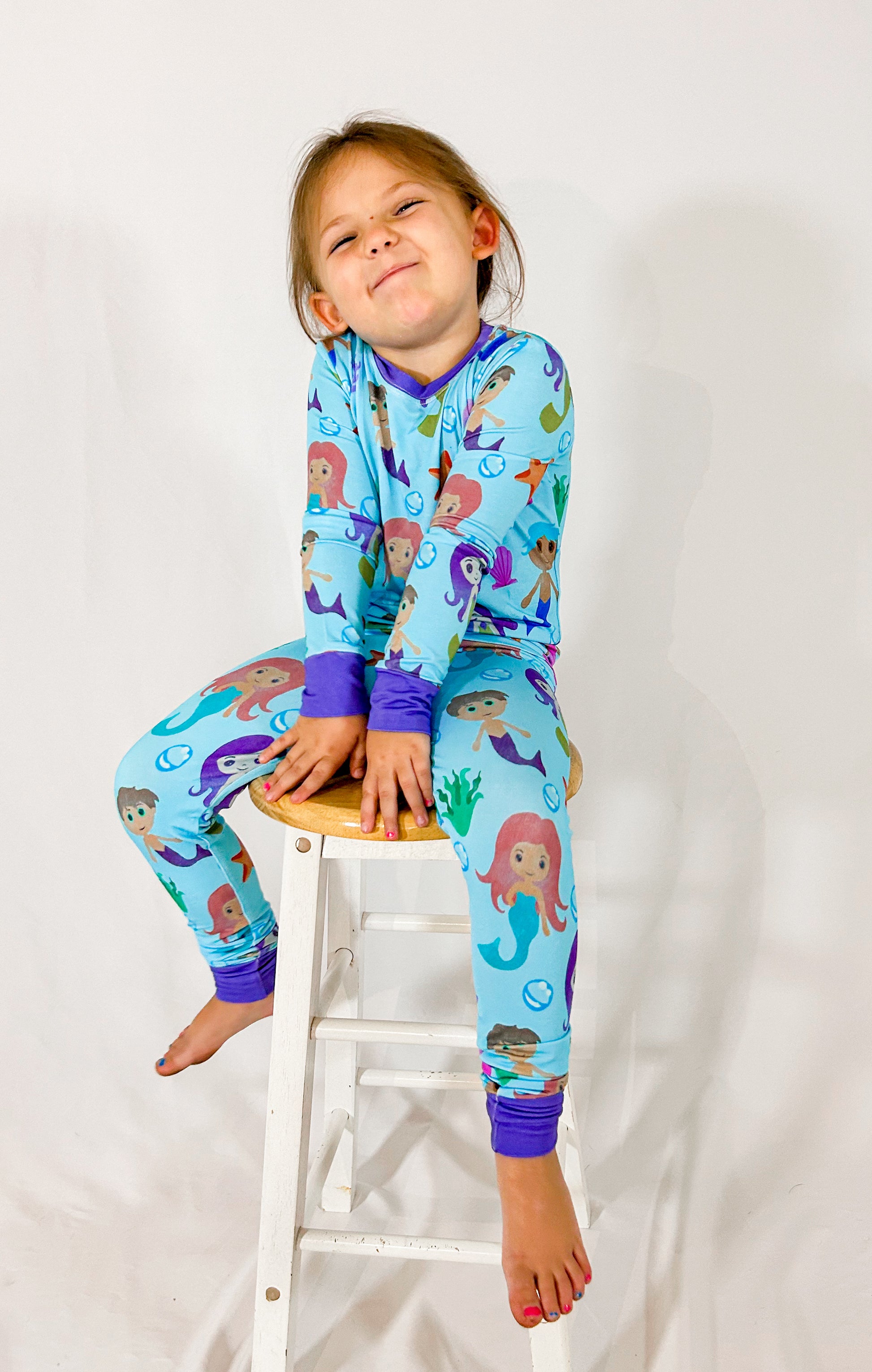 a girl sitting on a wooden stool wearing watercolor mermaid bamboo two piece pajama set