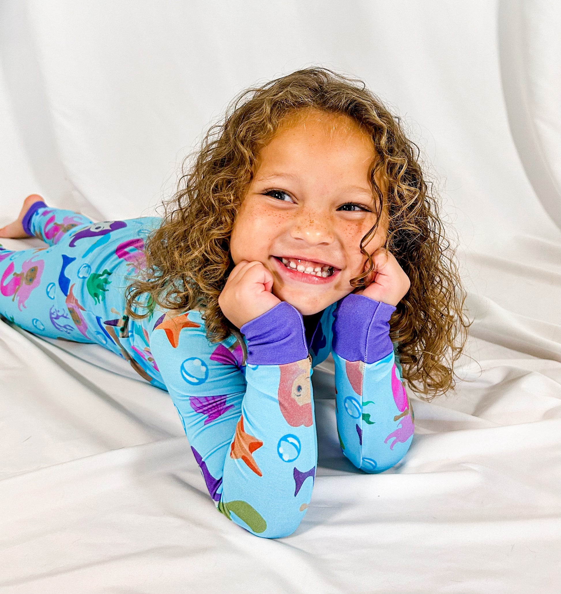 a smiling girl laying on the floor with her hands to her chin and wearing watercolor mermaid bamboo pajamas