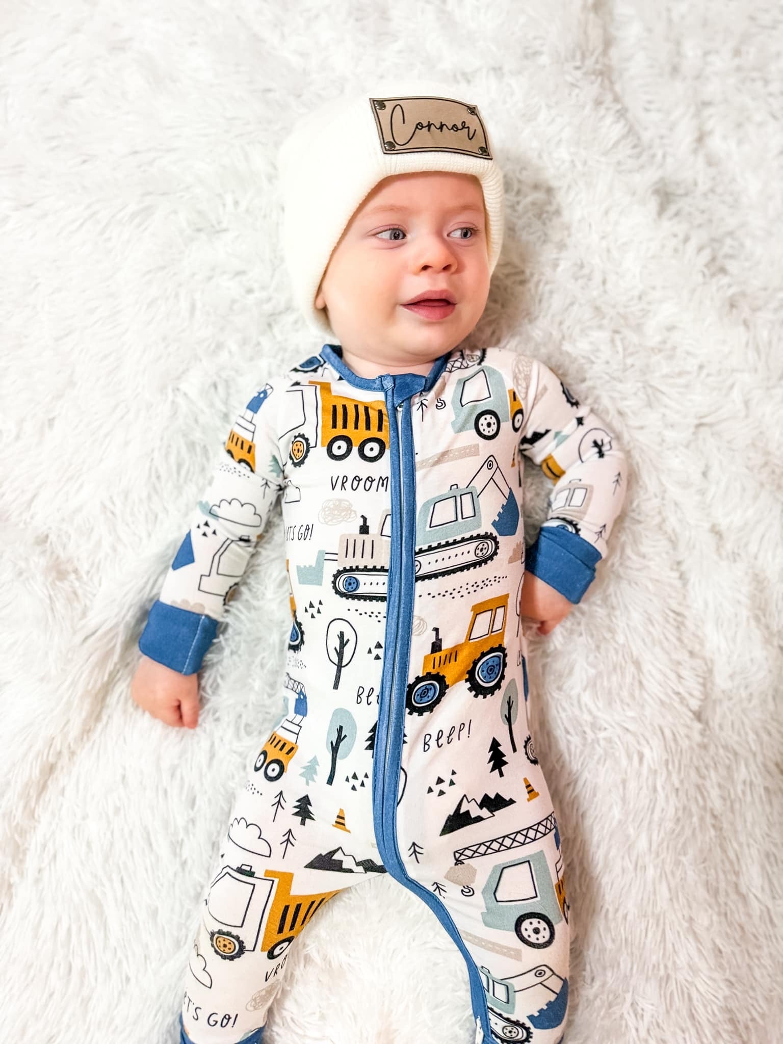 a young boy wearing a winter hat and laying on a fluffy white rug while wearing construction zone bamboo zippy featured excavators and mountains
