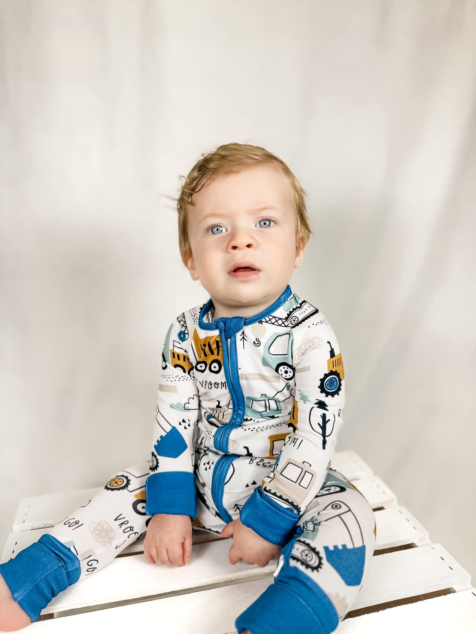 a young boy with bright blue eyes sitting on a wooden box wearing a construction zone bamboo zippy pajamas print
