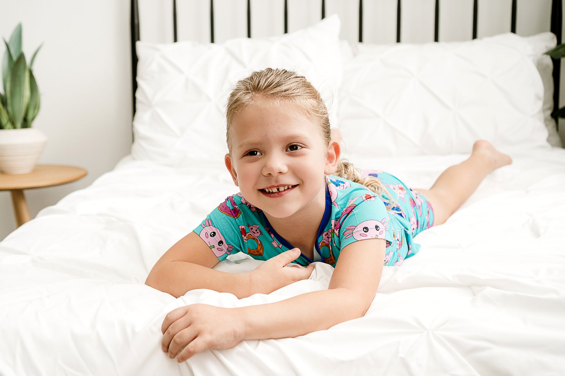 Girl lying on the bed wearing Axolotlicious bamboo shortie pajamas with a big smile on her face.
