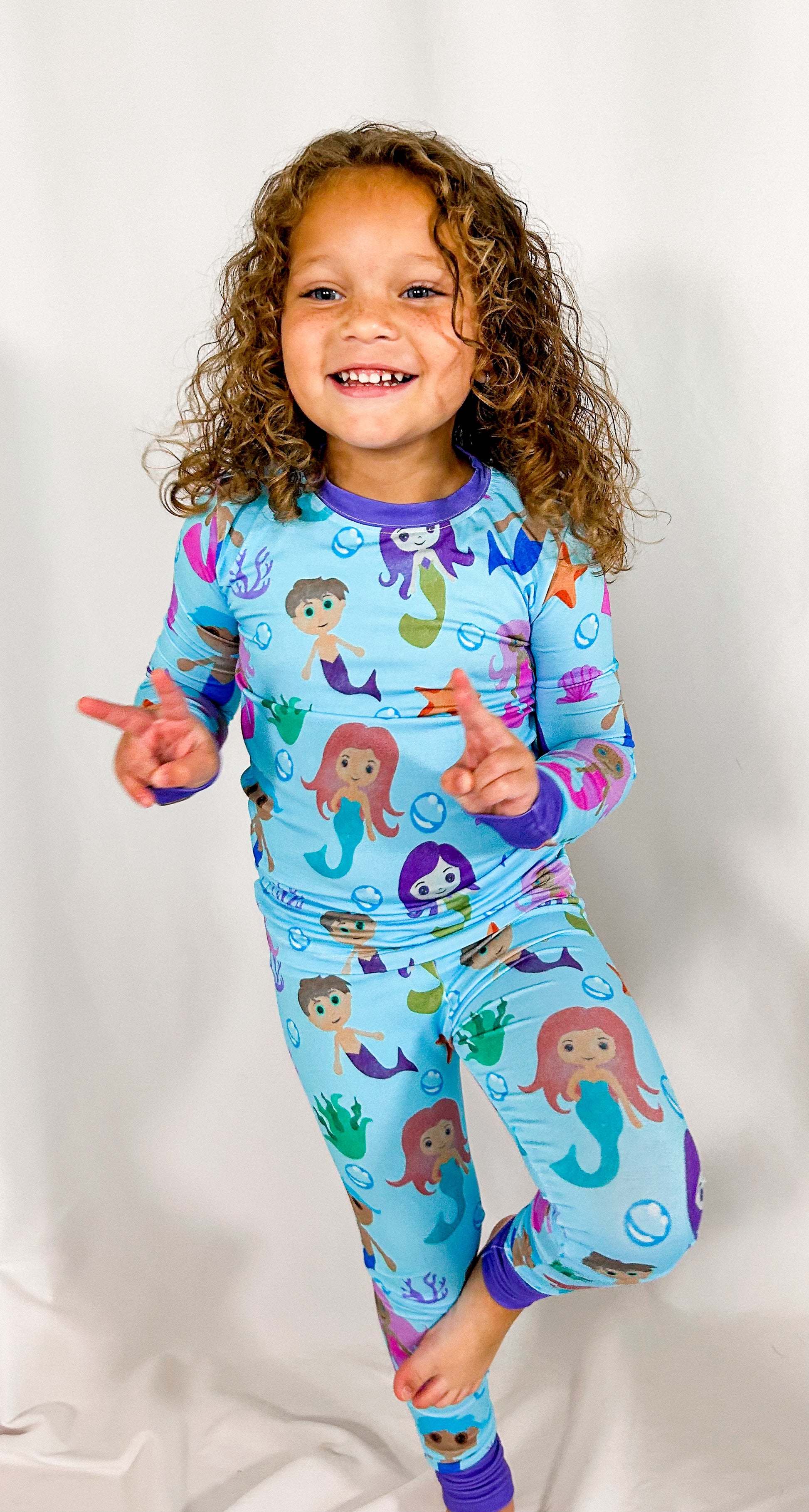 a girl standing on one foot and flashing peace signs while wearing watercolor mermaid bamboo two piece pajama set