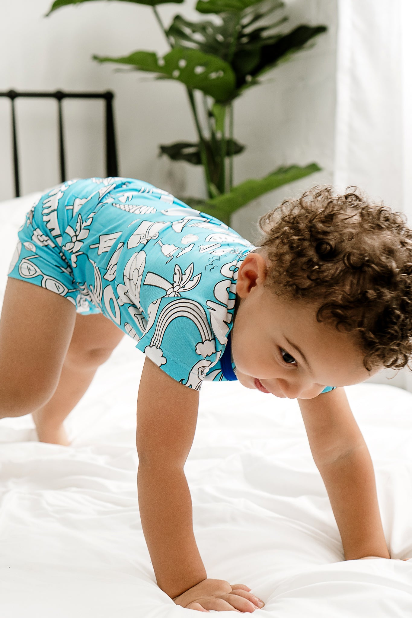 A child wearing a short sleeve/shorts pajama set with a beach and sea-themed print, including flip-flops, suns, and surfboards, climbing on a bed.
