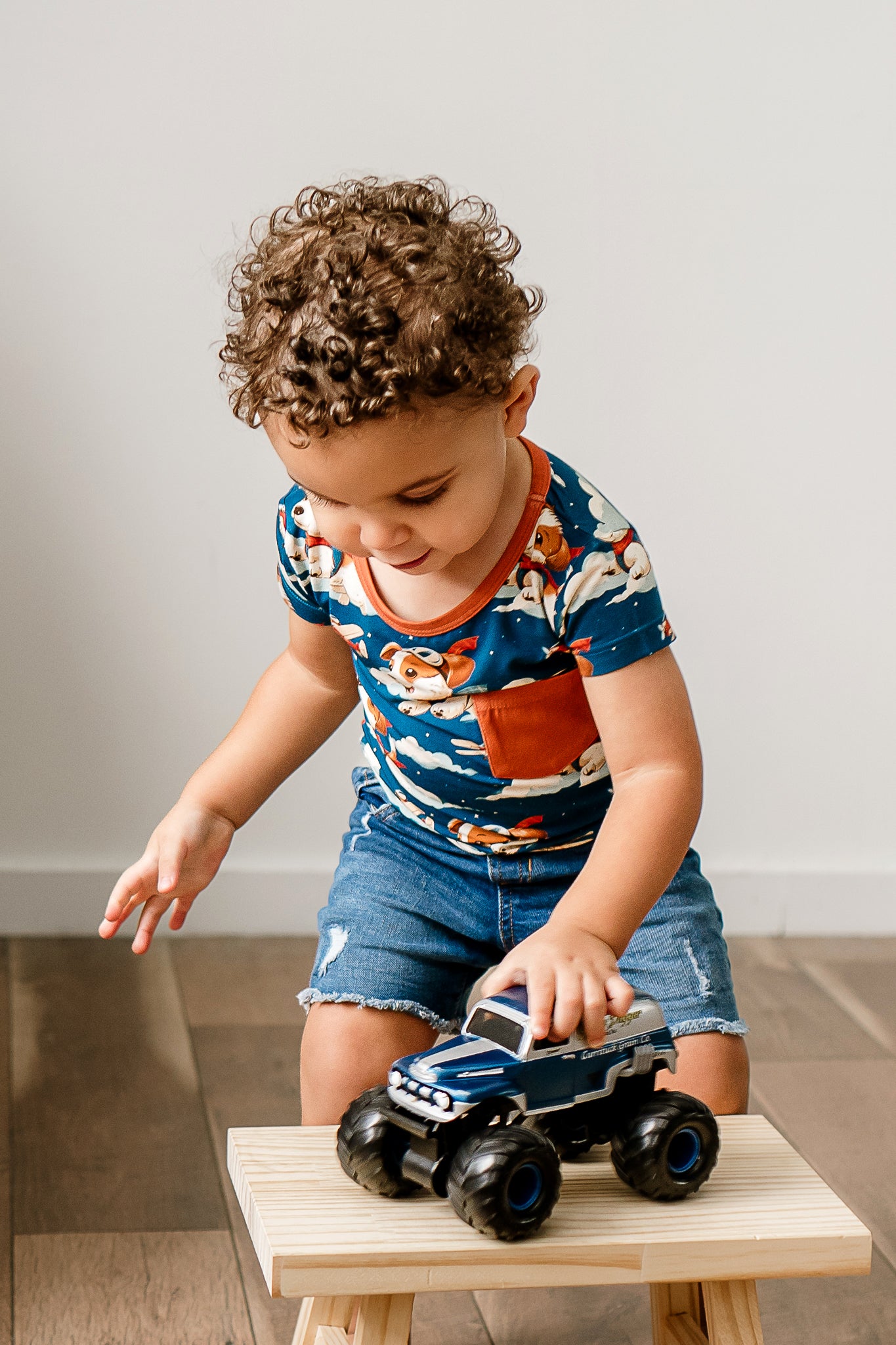 A child wearing a short-sleeve navy t-shirt with a print of dogs piloting airplanes called Paw-some Pilot, paired with denim shorts, sitting on a small wooden stool, playing with a toy car.