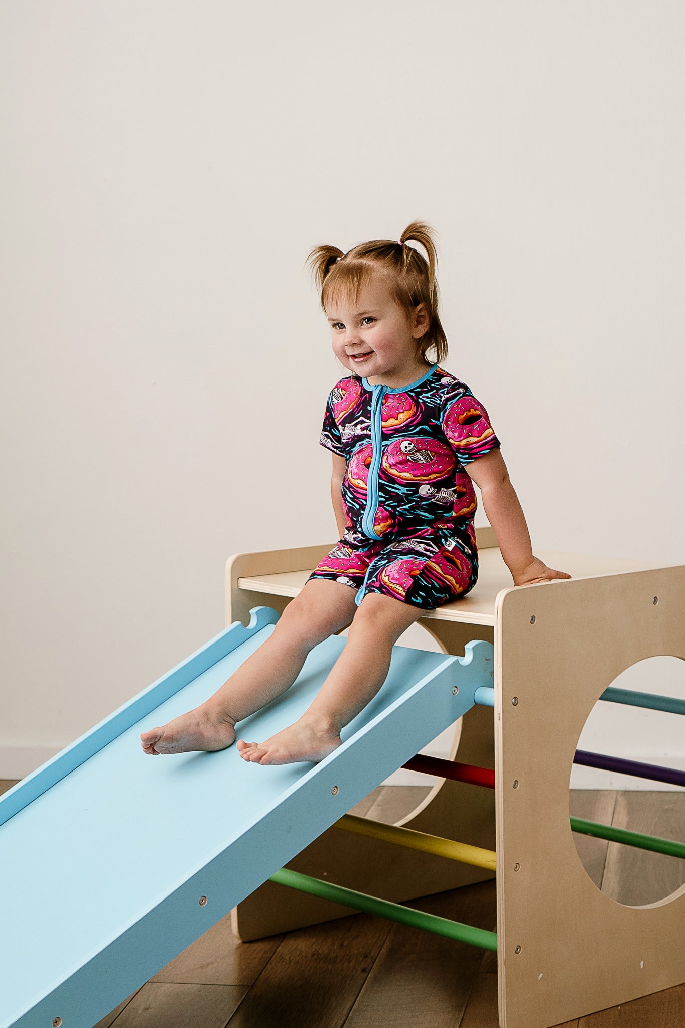 a girl smiling while sitting on the top of a wooden slide wearing ghoulish glaze bamboo zippy featuring donuts and skeletons