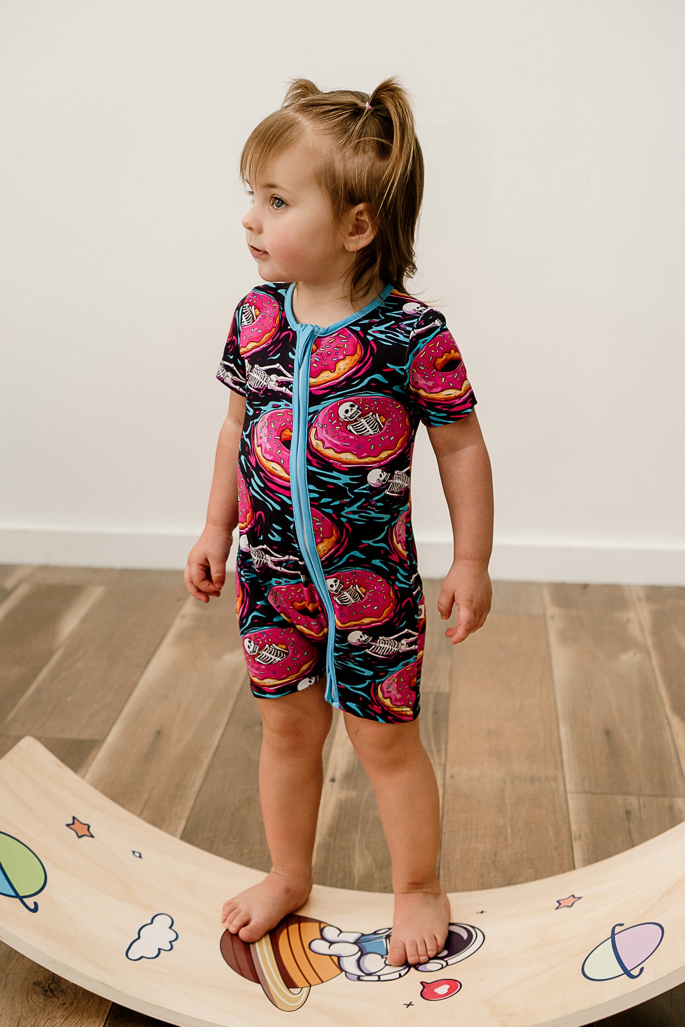 A child standing on a wooden floor wearing a multicolored short romper with a pattern of floating skeletons in donuts.