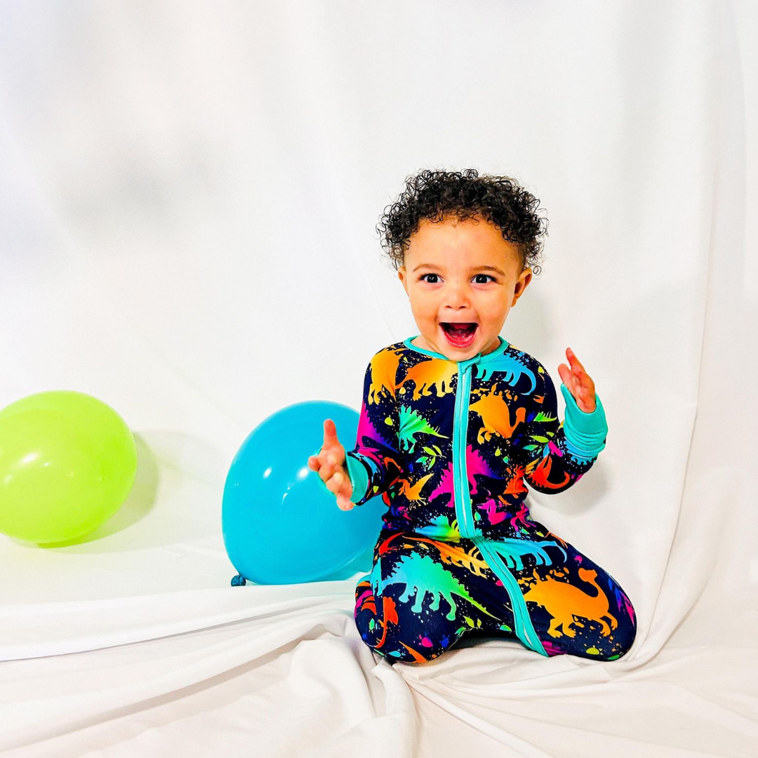 A child sitting on a white surface wearing multicolored dinosaur print pajamas with a prominent blue dinosaur on the front.