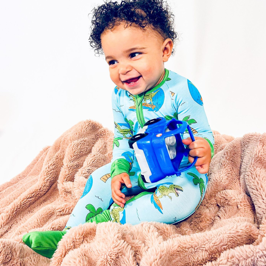 A toddler wearing blue pajamas with dinosaur and tropical print sitting on a beige blanket, holding a blue toy.