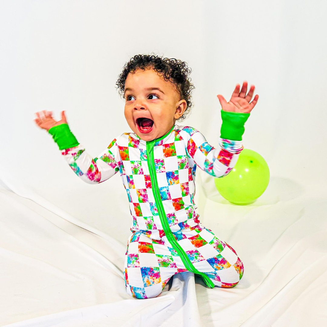 A child wearing a long-sleeved pajama set with a vibrant multicolored checkered pattern and green trim, sitting next to a green balloon.
