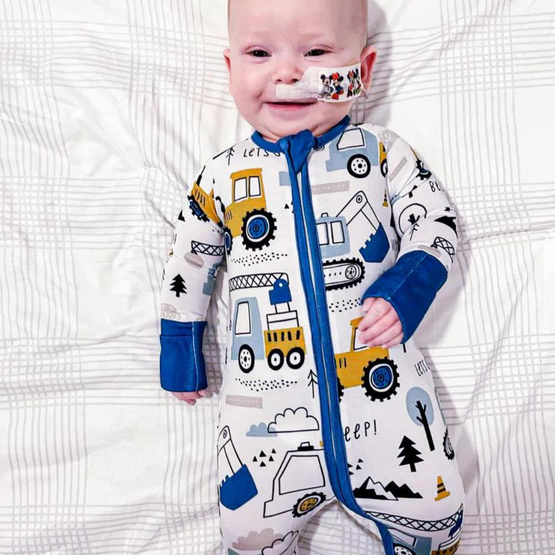 A baby wearing construction-themed pajamas with vehicles, trees, and mountains, lying on a striped surface.