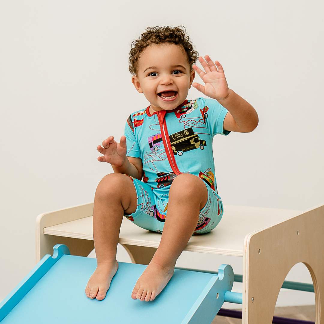 A baby sitting on a toy train set, wearing a short zippy with a colorful food print called Foodie Fun