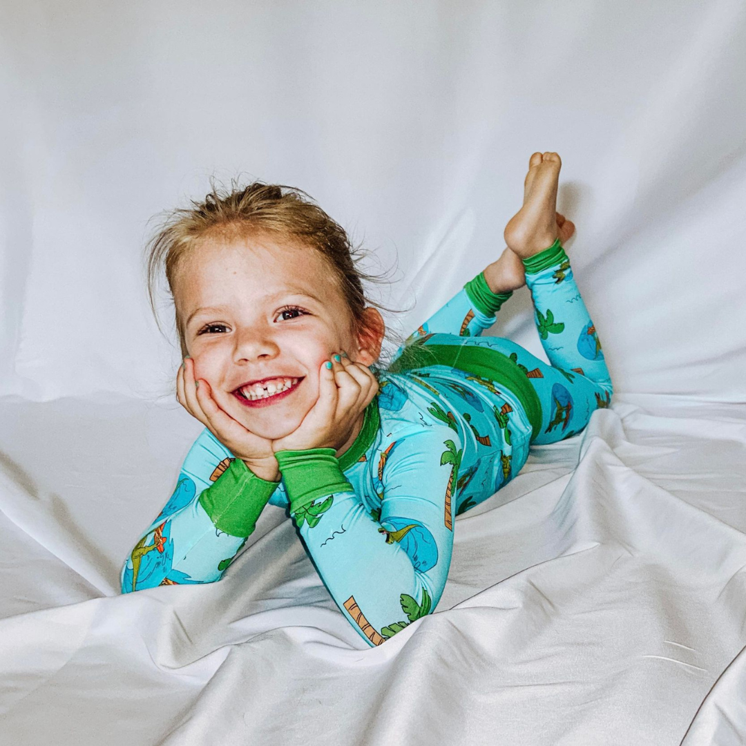 A child lying down wearing a two-piece pajama set with a tropical theme, including palm trees and surfing dinosaurs.