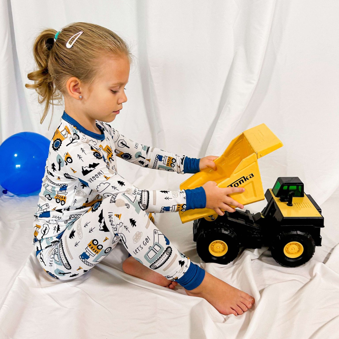 A child wearing a two-piece pajama set with construction theme, including trucks and building sites, sitting next to a toy dump truck.