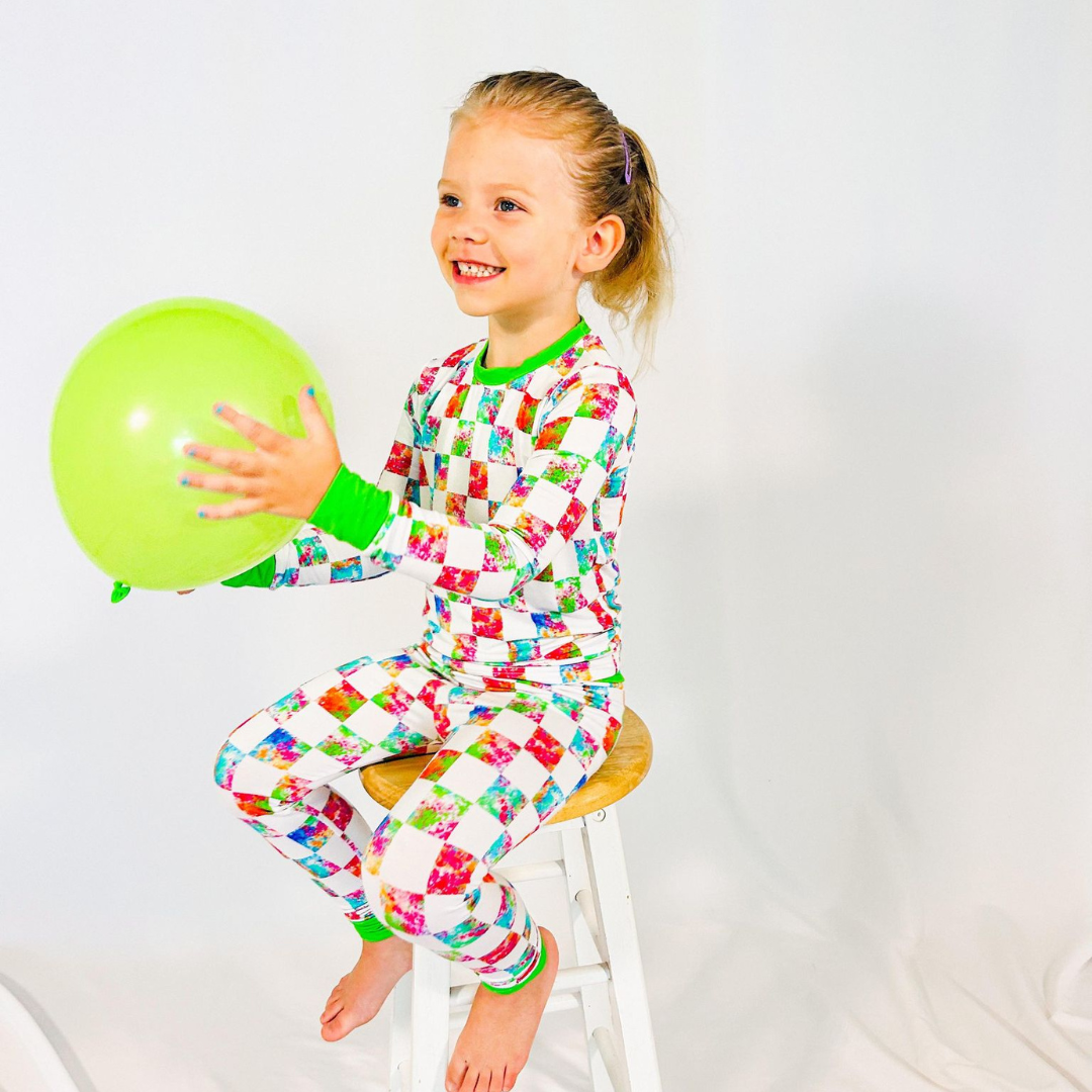 A child sitting on a stool wearing a colorful checkered pajama set with long sleeves and full-length pants, holding a green balloon.