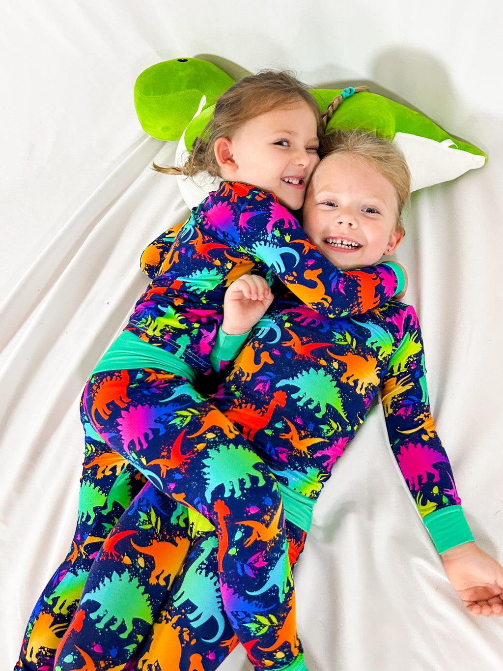 two girls giving eachother a playful hug while laying their head on a dinosaur stuffy and wearing matching neon dinosaur pajamas