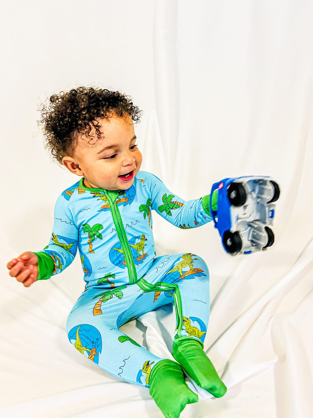 a boy sitting on the floor playing with a toy car while wearing a surfing dinosaurs print bamboo zippy with fold over feet