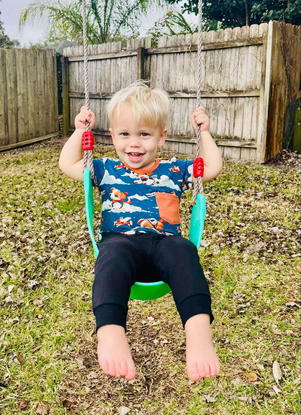 A child sitting on a swing set in an outdoor setting, wearing a short sleeve shirt with a pilot theme.