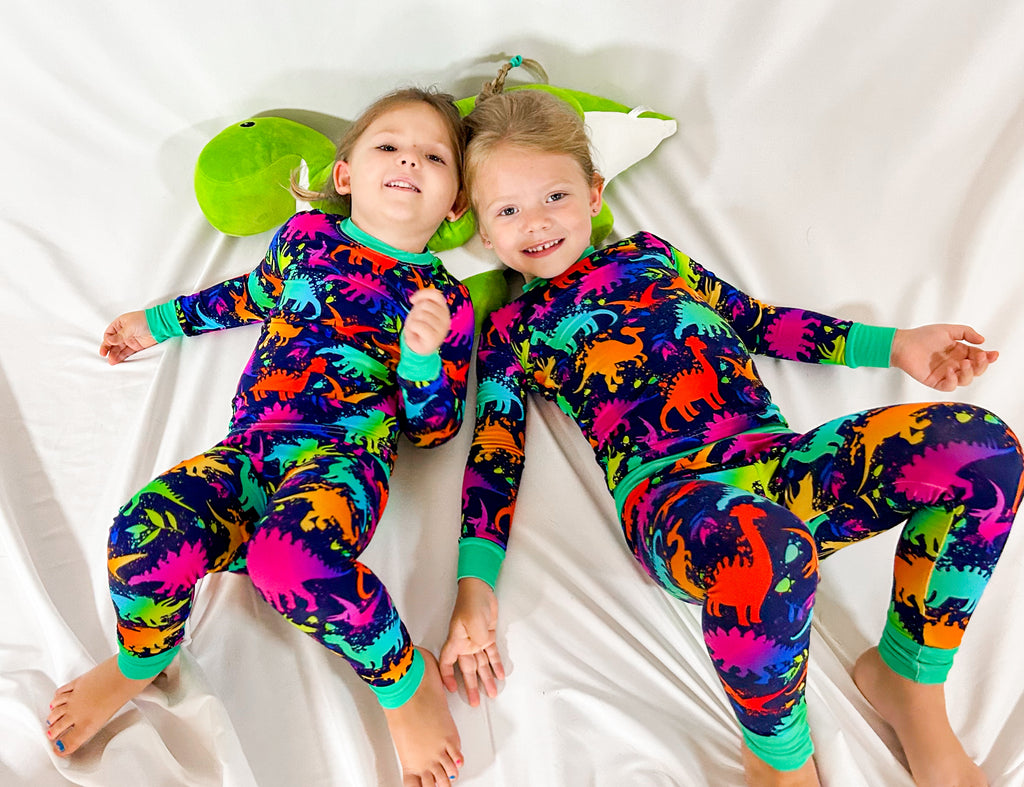 Two children lying down wearing two-piece pajama sets with vibrant dinosaur prints on a colorful background.