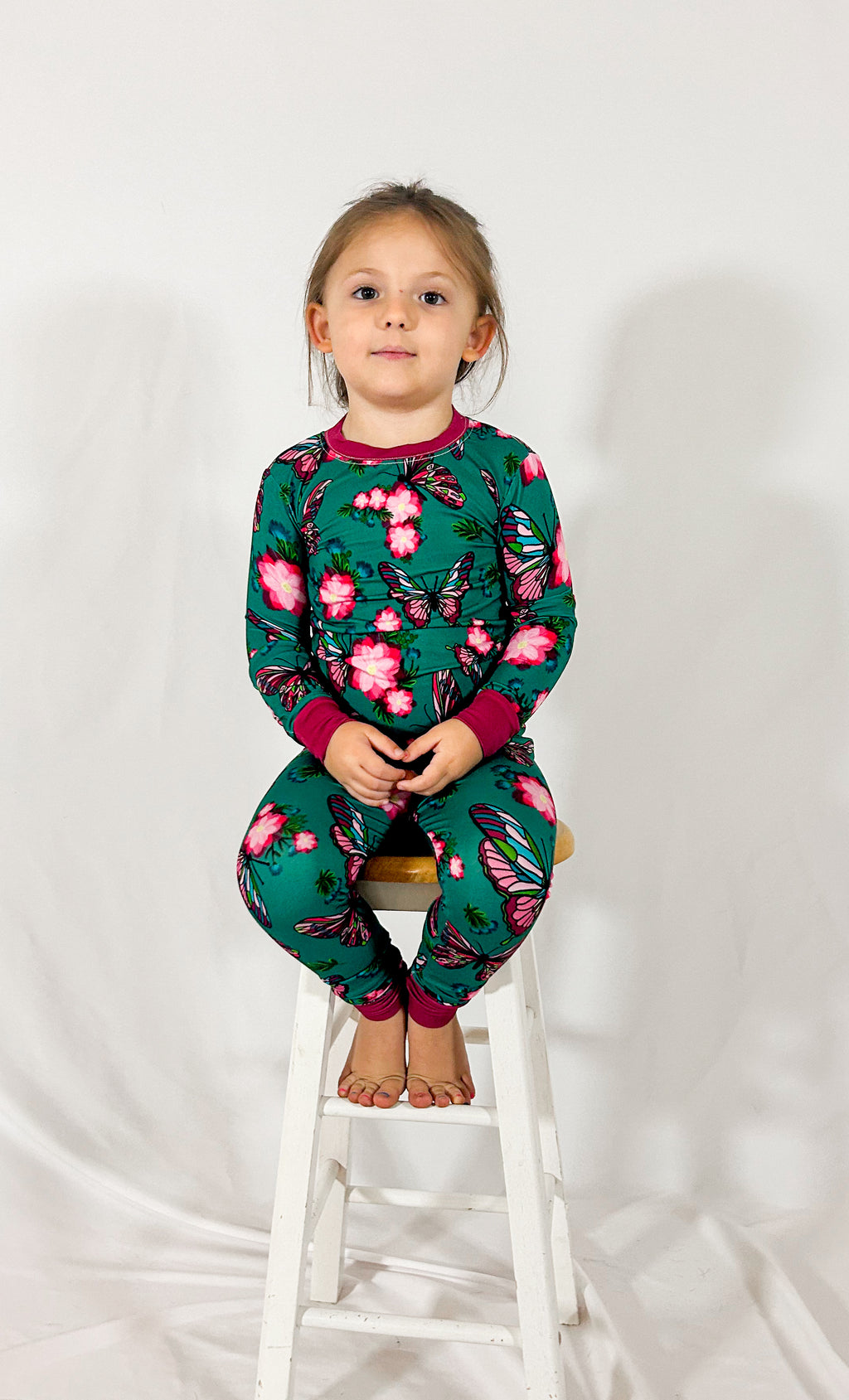 a girl sitting on a wooden stool wearing our emerald butterfly bamboo pajamas in an emerald green color 