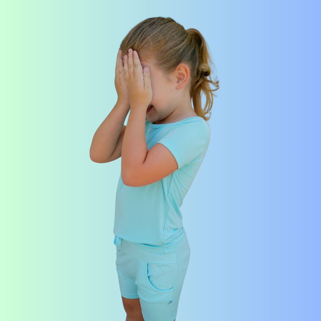 A little girl wearing Arctic Blue Bamboo Shirt and Shorts playfully covering her eyes on a blue gradient background