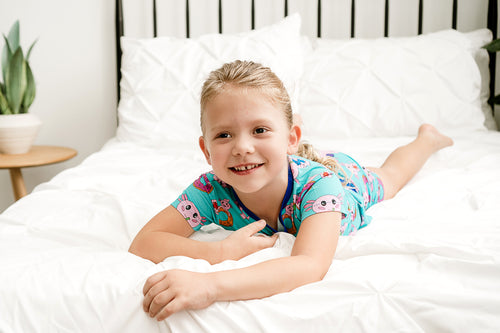 Girl lying on the bed wearing Axolotlicious bamboo shortie pajamas with a big smile on her face. 