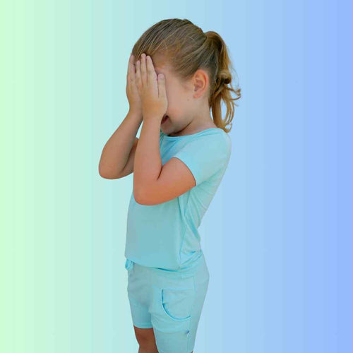 Young child in breathable Arctic blue bamboo shorts, playfully covering her eyes, showcasing flexible fit and comfort.