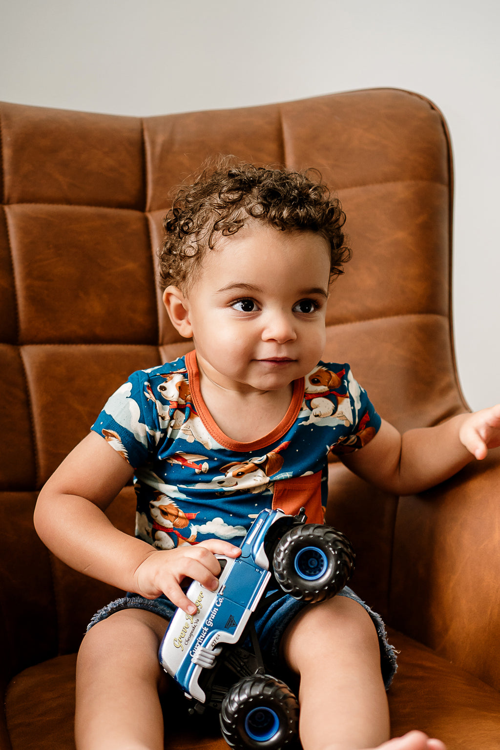 a boy sitting in a brown leather chair, holding a monster truck toy and wearing Paw-some Pilot bamboo shirt