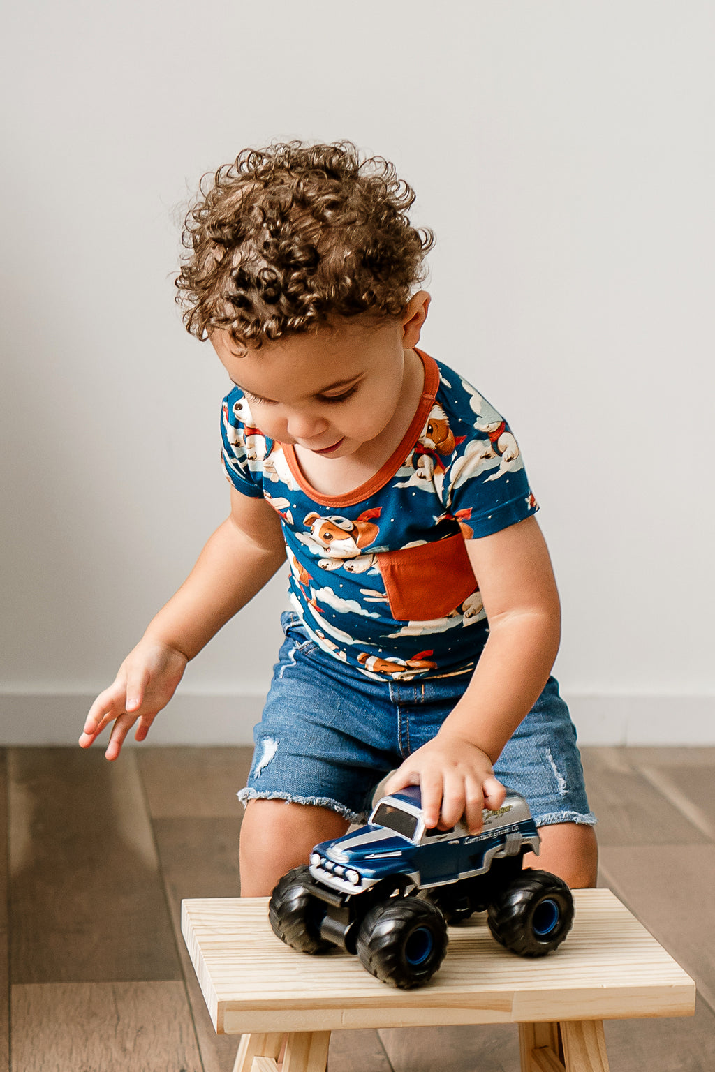 A child wearing a short-sleeve navy t-shirt with a print of dogs piloting airplanes called Paw-some Pilot, paired with denim shorts, sitting on a small wooden stool, playing with a toy car.