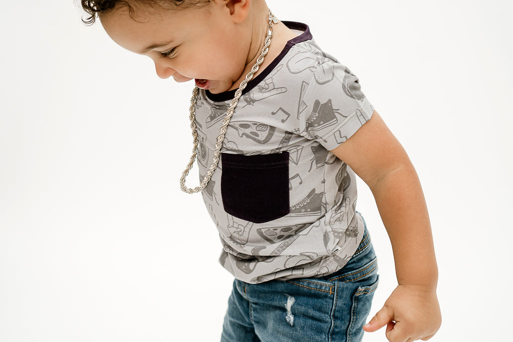 A child wearing a short sleeve shirt with a pattern of skulls, guitars, and pizza slices against a grey background called Tiny Rebel