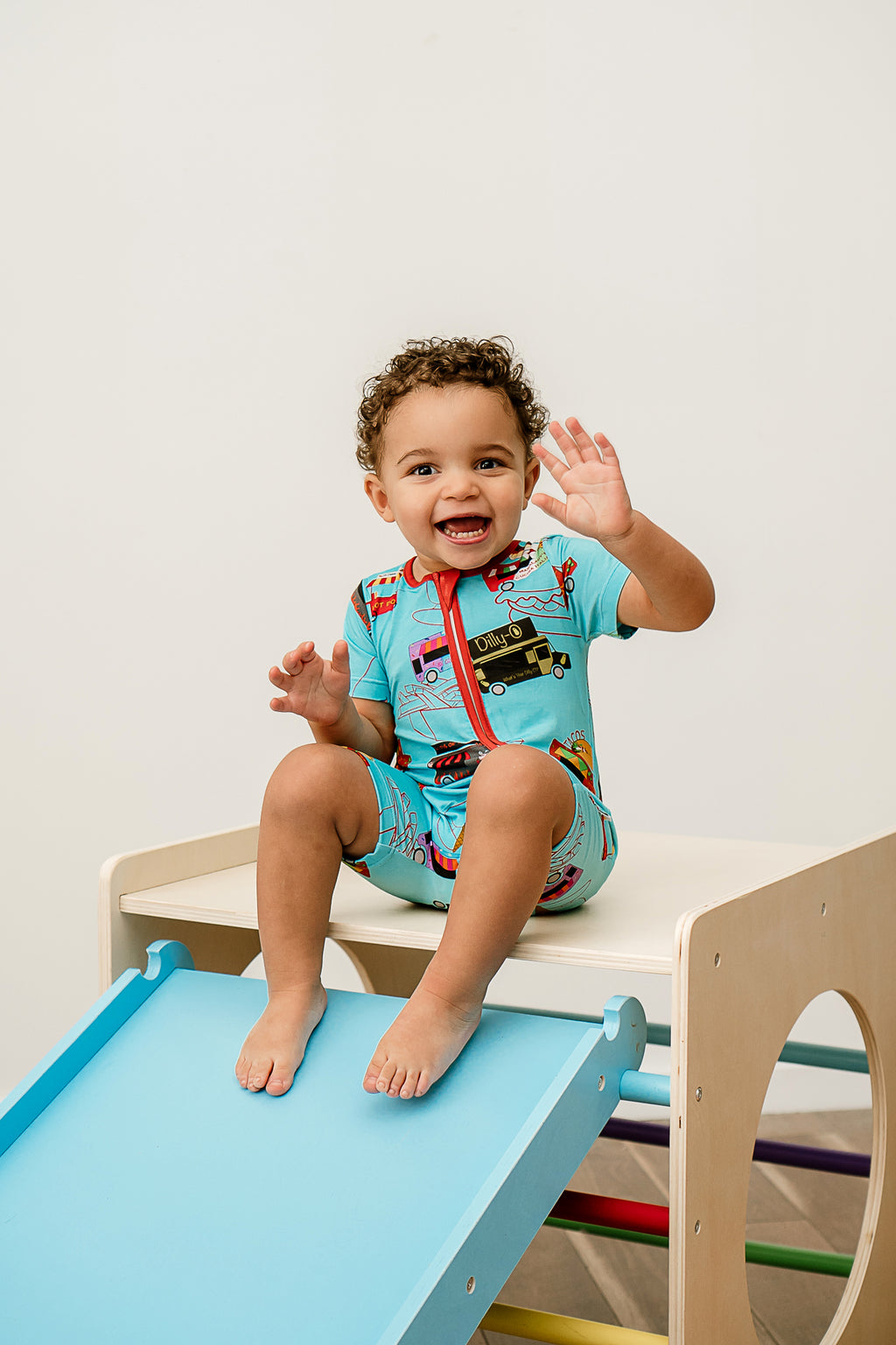 a joyful boy smiling and waving sitting on a wooden box wearing Foodie Fun Bamboo Zippy