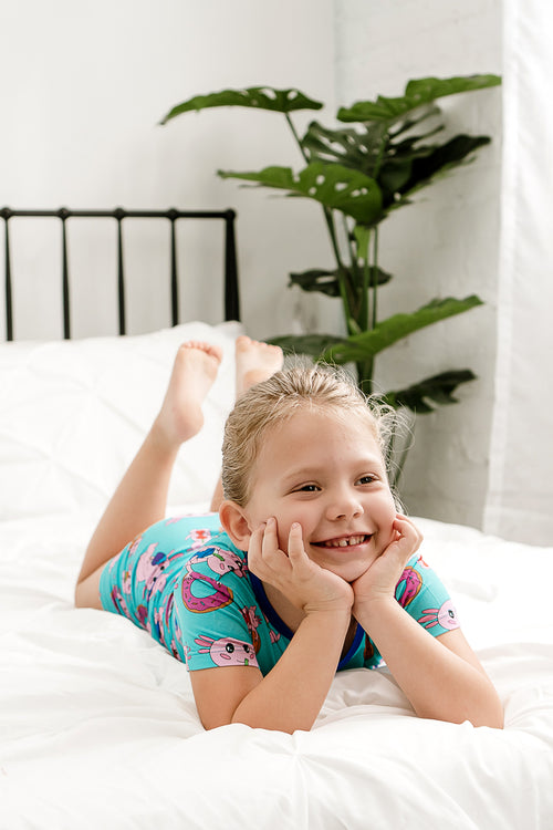 Girl relaxing on a bed wearing Axolotlicious bamboo short sleeve zippy with axolotl print, smiling peacefully.