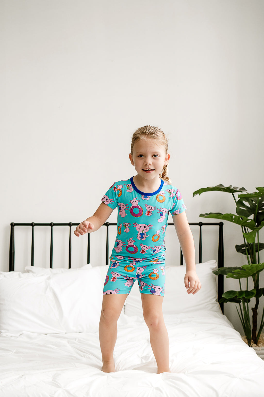 Girl smiling and jumping on the bed in Axolotlicious bamboo pajamas with short sleeves and shorts.