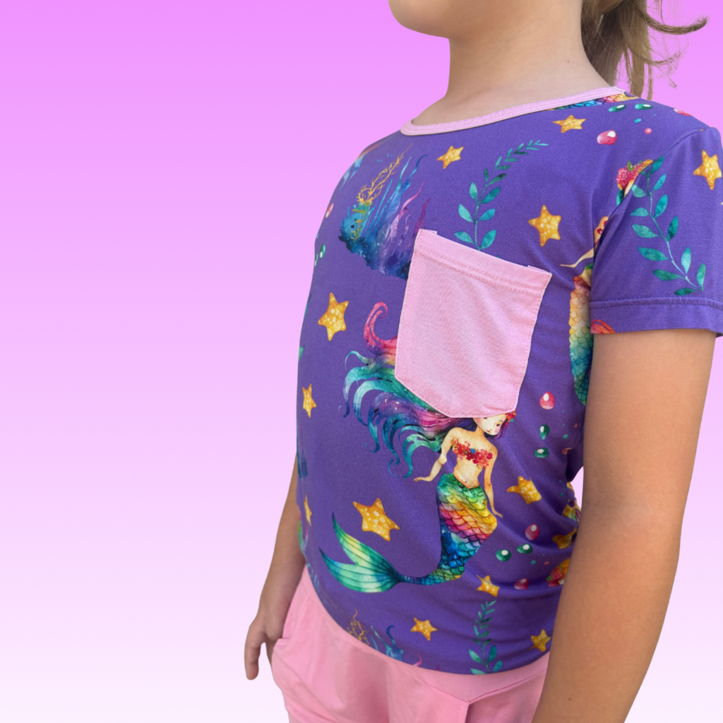 Toddler girl in a soft, breathable Rainbow Reef bamboo shirt featuring vibrant ocean-inspired colors, with Pink Lemonade Bamboo shorts and a purple gradient background