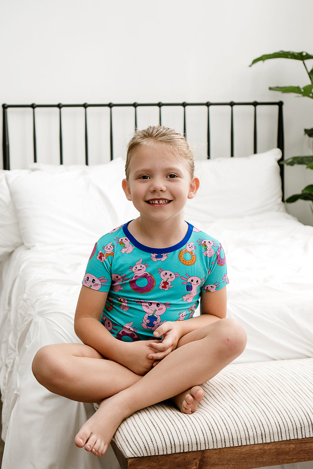 Happy child sitting on the foot of the bed while dressed in vibrant Axolotlicious bamboo PJs.
