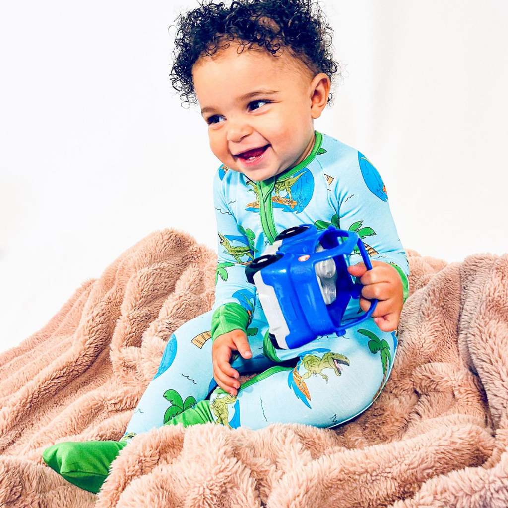 A toddler wearing blue pajamas with dinosaur and tropical print sitting on a beige blanket, holding a blue toy.