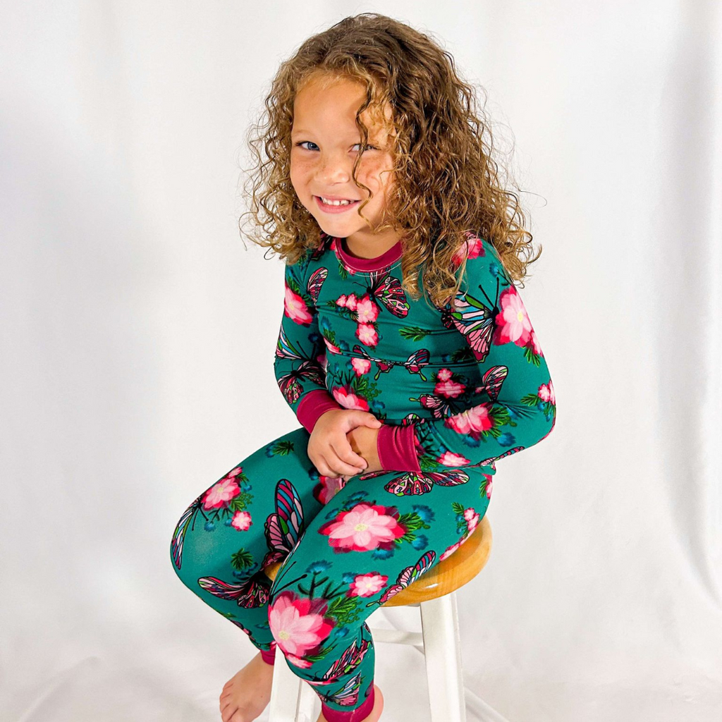 A child sitting on a stool wearing a two-piece pajama set with a green base and pink floral print, including butterflies, and burgundy trim.