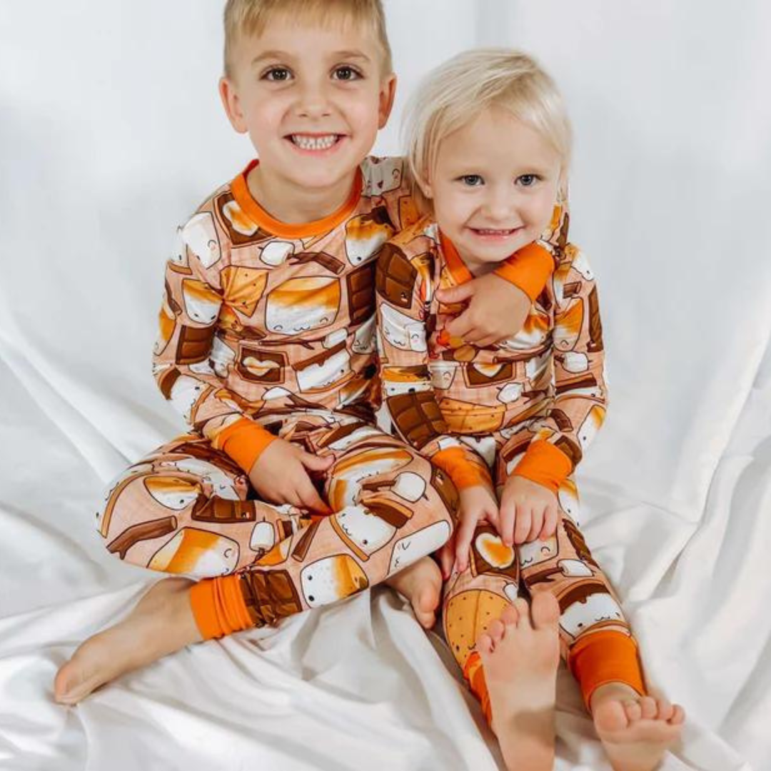 Two children wearing orange and brown s'mores-themed pajamas, sitting on a white surface.