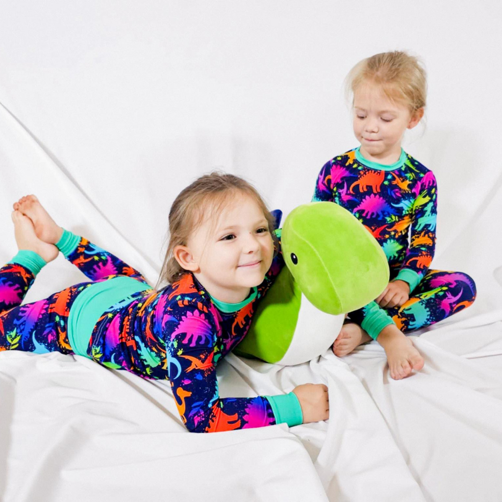two girls wearing matching neon dinosaur bamboo pajamas and cuddling with a dinosaur stuffy